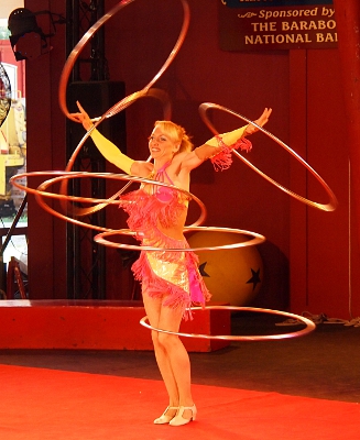 [Woman in yellow costume with long pink fringe twirling 2 hula hoops on one hand 1 hoop on another, 2 around her waist and 1 at her knees.]